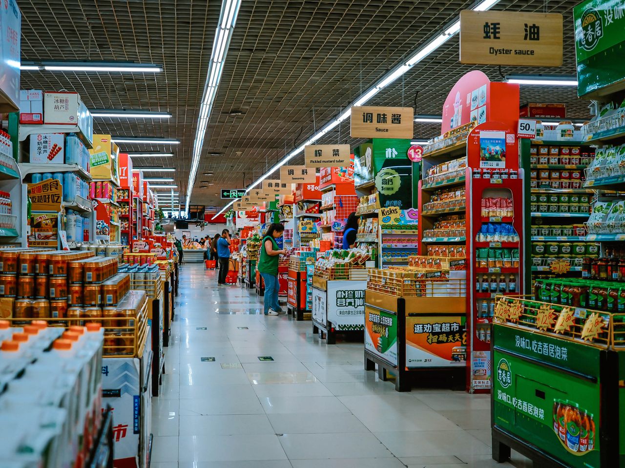 gallery-4 A vibrant supermarket aisle filled with diverse products and shoppers under bright lighting.